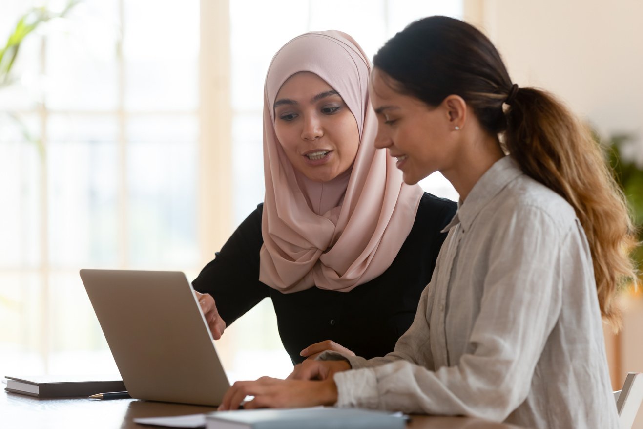 Two individuals in hijab sitting at a table looking at a laptop. One of them is a food safety consultant , helping the food factory owner how Food Audit & mentoring are important for her food business operations.