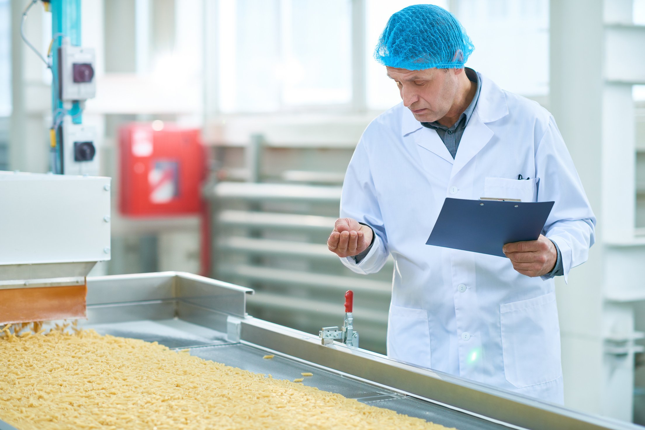 factory supervisor in a lab coat is looking at food products on a conveyor belt being manufactured in his factory, while he is considering if all requirements relating to the HACCP is being met.