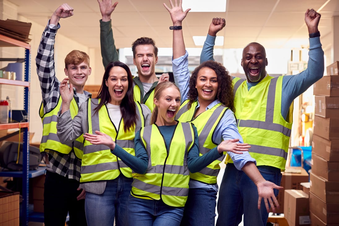 a group of employees wearing safety vests in a warehouse, celebrating successful completion of their health & safety training at work.