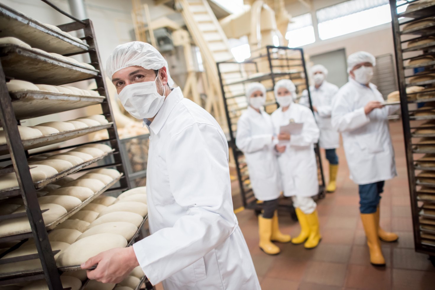 factory workers in white lab coats with PPE are working in a bakery, making bread. there are also food safety auditors and quality controls having a word.